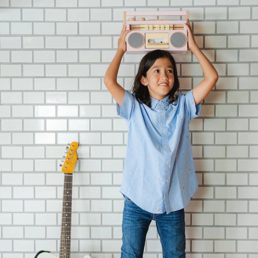 A young girl holding a Retro Wooden Tape Recorder above her head in a room with a white tiled wall and a yellow electric guitar leaning nearby. She smiles and looks upwards, wearing a blue shirt.