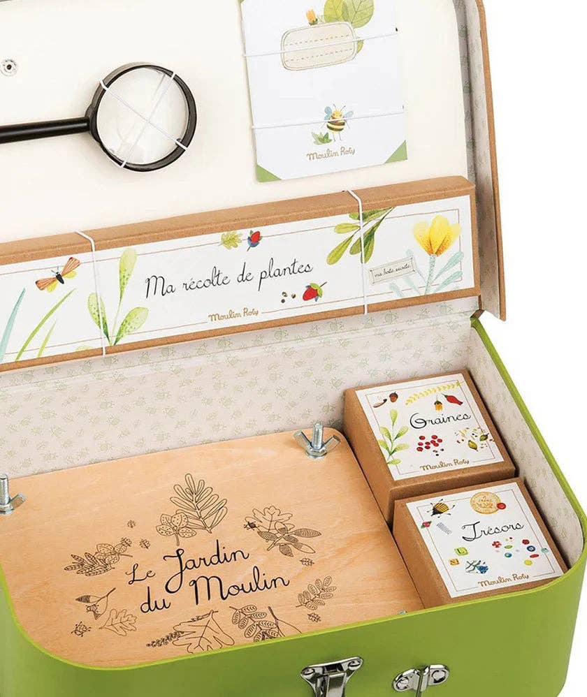 The Moulin Roty Botanist Suitcase is a green suitcase-style box for young botanists, with gardening-themed items inside—illustrated cards, a magnifying glass, wooden compartments, and labeled "Graines" and "Trésors" boxes.