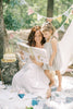 A woman in a white dress reads Winnie-the-Pooh: Finding Feelings to a young girl outdoors. They sit on a blanket by a tree, surrounded by cake and bunting for a cheerful, festive scene.