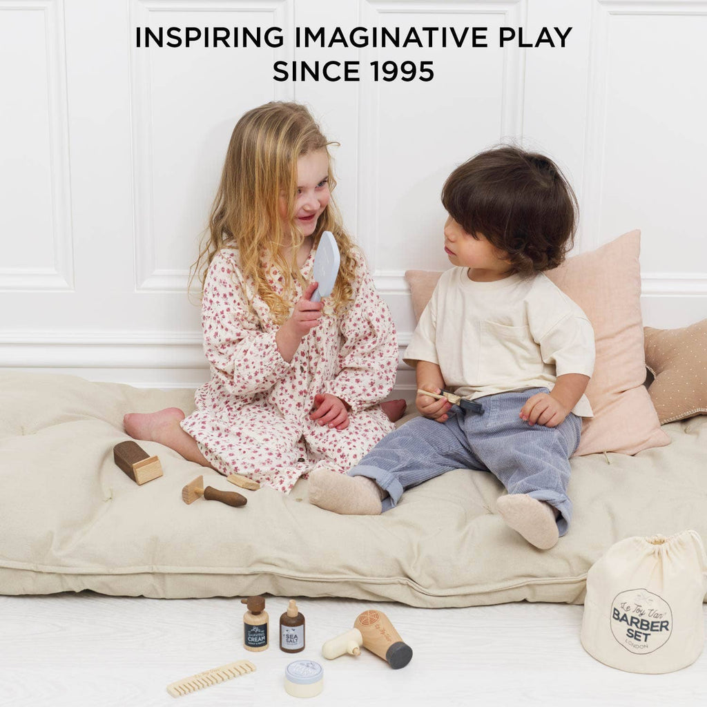 Two young children sit on a cushion playing with the Wooden Hairdresser & Barber Kit, smiling at each other. Above them: "Inspiring Imaginative Play Since 1995." The plastic-free kit and small bag are on the floor. Ships in one week.