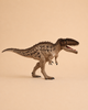 The Schleich Carcharodontosaurus Dinosaur Toy, featuring a brown textured body with black markings, stands on two legs against a plain beige background with its mouth open to reveal small teeth.