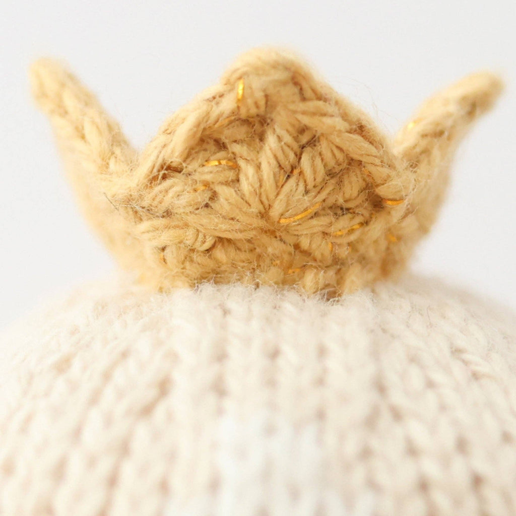 A close-up of Cuddle + Kind Tiny Rose the Princess, a small, handmade knitted crown with gold threads from Peru, resting on a cream knit surface against a plain light background.