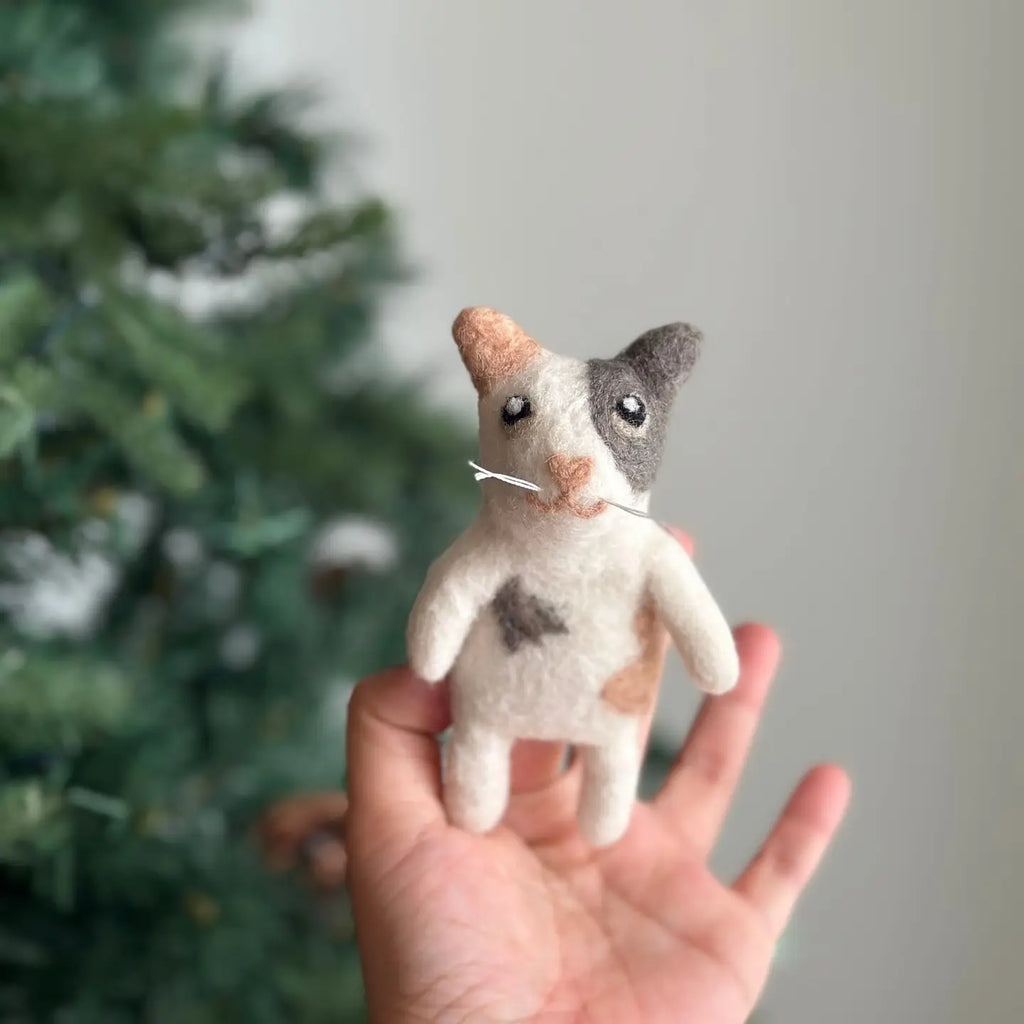 The Calico Cat Finger Puppet, a handmade wool creation, is being displayed against a blurred background with a Christmas tree. This felted puppet features patches of black, white, and brown, and includes a small, delicate nose and whiskers, making it an ideal whimsical storytelling prop.