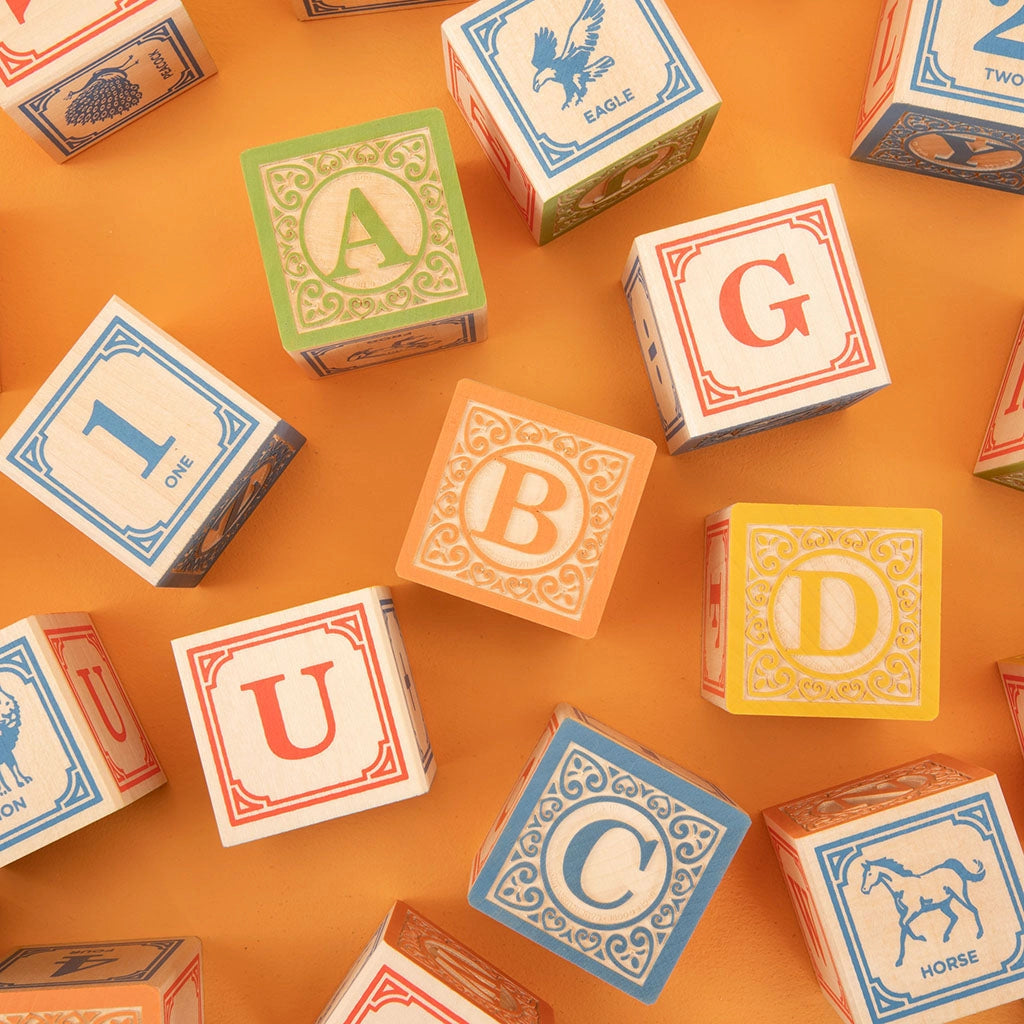 Sentence with Product Name: Uncle Goose Classic ABC Blocks scattered on an orange background, featuring letters and illustrations such as animals and numbers.
