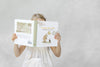 A young girl in a white dress sits against a light gray background, holding and reading Winnie-the-Pooh: Finding Feelings, her face mostly hidden behind the cover.