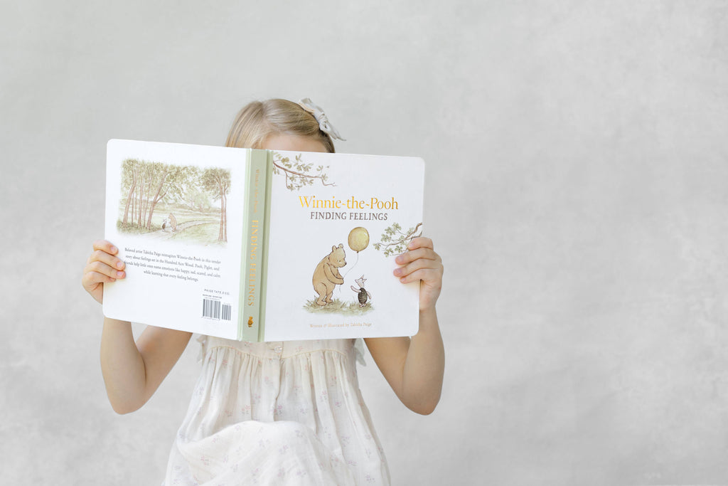 A young girl in a white dress sits against a light gray background, holding and reading Winnie-the-Pooh: Finding Feelings, her face mostly hidden behind the cover.