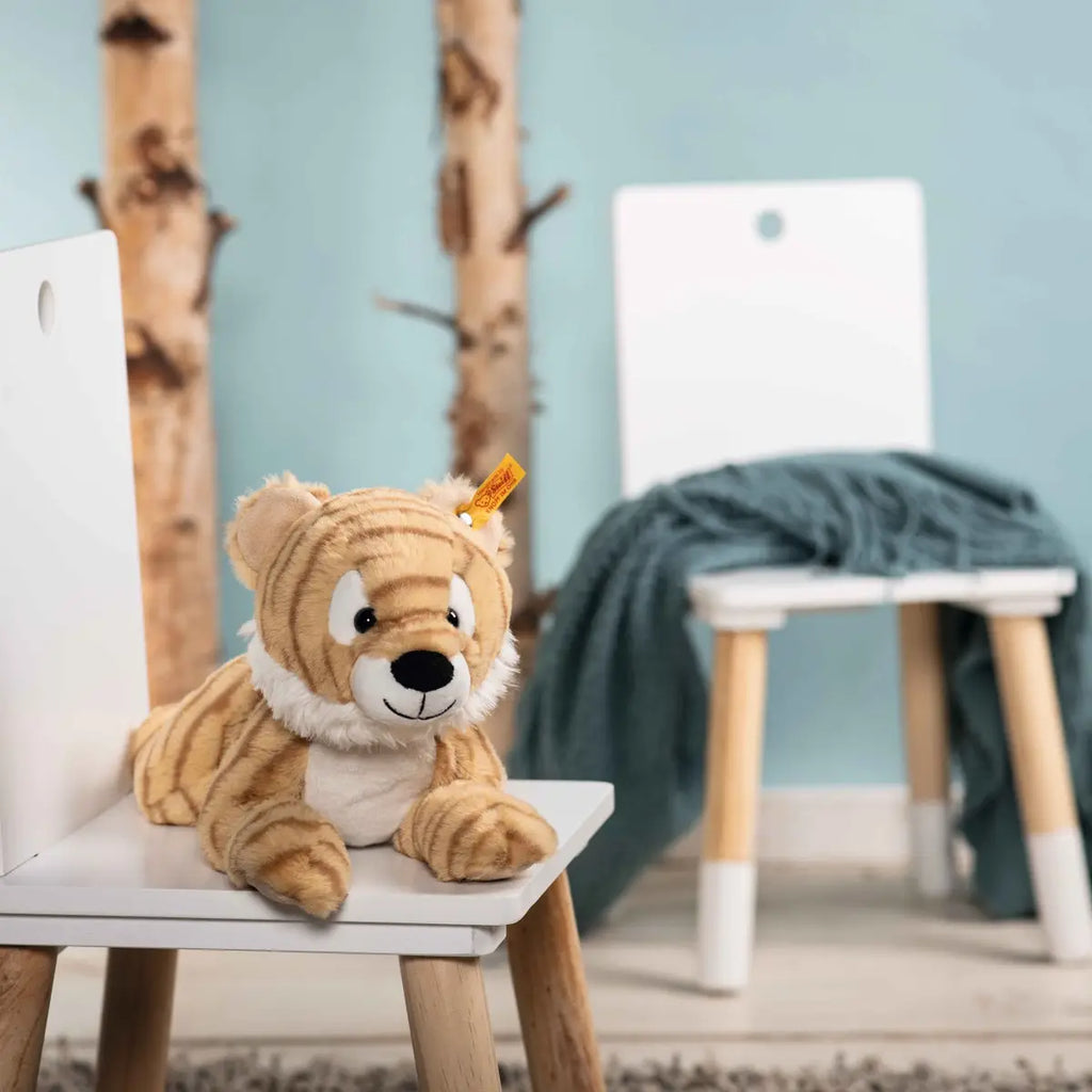 A Steiff, Toni Tiger Stuffed Animal Plush, 12 Inches with a friendly expression is lying on a white chair. Another similar white chair with a blue blanket draped over it is in the background. The chairs are wooden, and the room has nature-inspired decor with tree trunks against a light blue wall—perfect for Soft Cuddly Friends.