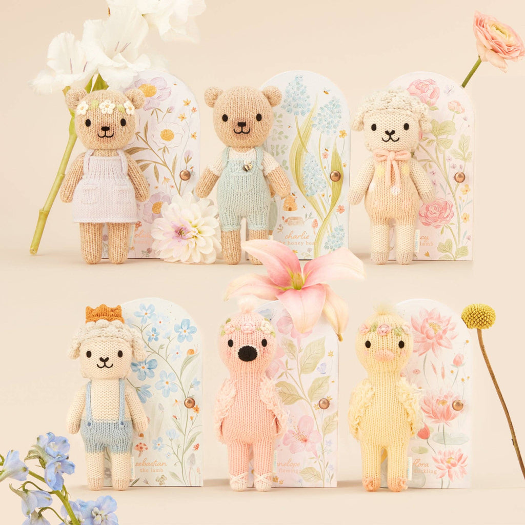 Cuddle + Kind Tiny Charlie the Honey Bear, a fair trade hand-knit doll handmade in Peru, stands before a pastel floral background wearing a soft-colored outfit and flower crown.