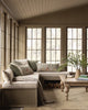 A cozy sunroom with large windows, a beige sectional bench featuring the Coral & Tusk Mushrooms and Ferns Lumbar Pillow among patterned and solid cushions, a wooden coffee table with books and greenery, plus firewood stacked beneath the bench.