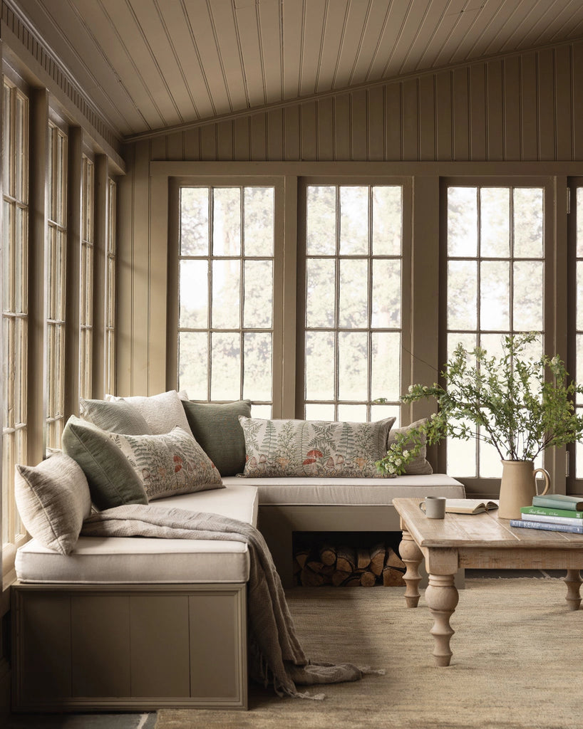 A cozy sunroom with large windows, a beige sectional bench featuring the Coral & Tusk Mushrooms and Ferns Lumbar Pillow among patterned and solid cushions, a wooden coffee table with books and greenery, plus firewood stacked beneath the bench.