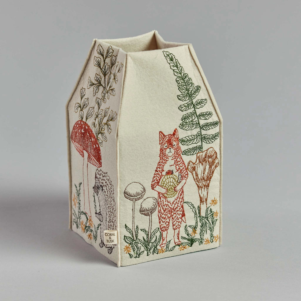 A Coral & Tusk Mushrooms and Ferns tissue box cover in beige, adorned with embroidered designs featuring a fox standing upright, various types of mushrooms, and plants, set on a plain grey background.