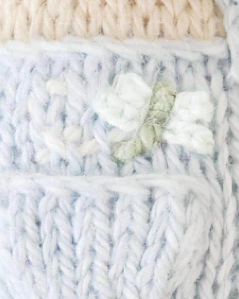 Close-up of Cuddle + Kind Tiny Bailey the Mouse, featuring light blue and cream knitted fabric, handmade in Peru, with a delicate embroidered white flower and green leaves.