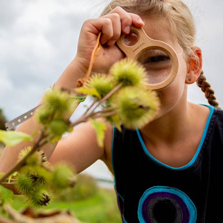A young girl with braided hair uses the Huckleberry Dual Magnifier to closely examine a spiky green plant outdoors, focusing intently on its details.