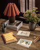 A wooden desk with notebooks, a Coral & Tusk Snowman Card, pencils, illustrated cards, a pen holder, magazines, a potted plant, and a brown lamp with a pleated shade creates cozy winter decor by the window with shutters.