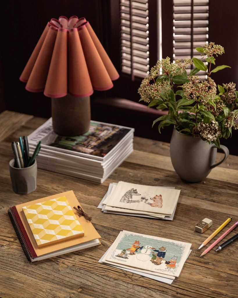A wooden desk with notebooks, a Coral & Tusk Snowman Card, pencils, illustrated cards, a pen holder, magazines, a potted plant, and a brown lamp with a pleated shade creates cozy winter decor by the window with shutters.