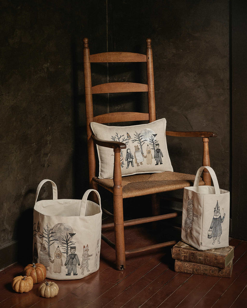 A wooden chair with the Coral & Tusk Full Moon Masquerade Pillow sits next to two whimsical animal fabric baskets, with small pumpkins and books on the floor against a dark wall.