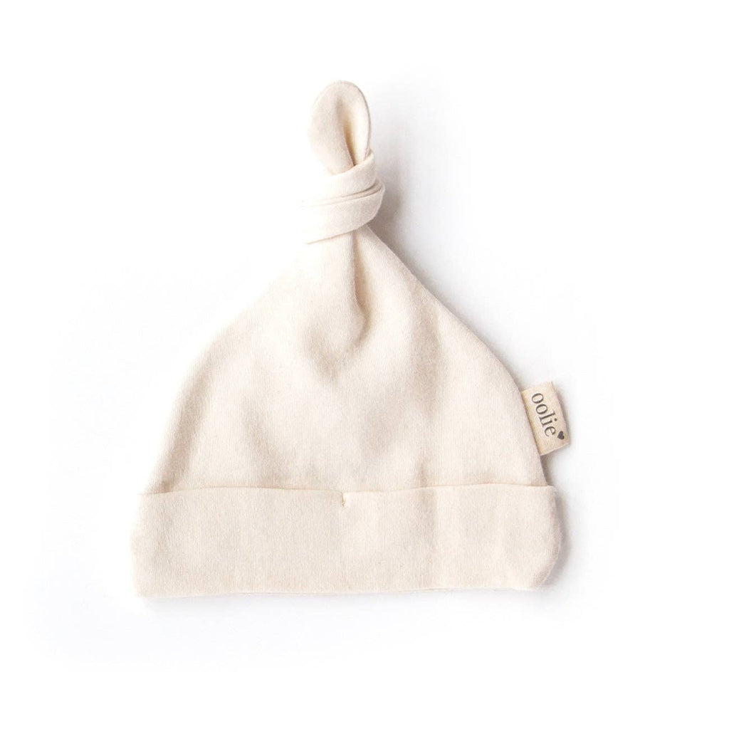 The Organic Baby Hat – Natural is a cream-colored, soft organic cotton hat for newborns with a folded brim, knotted top, and a small side tag, shown on a white background.