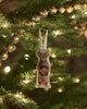 The Coral & Tusk Mouse with Wreath Ornament, dressed in red overalls and holding a wreath, hangs from a Christmas tree branch decorated with golden bead garland and glowing string lights.