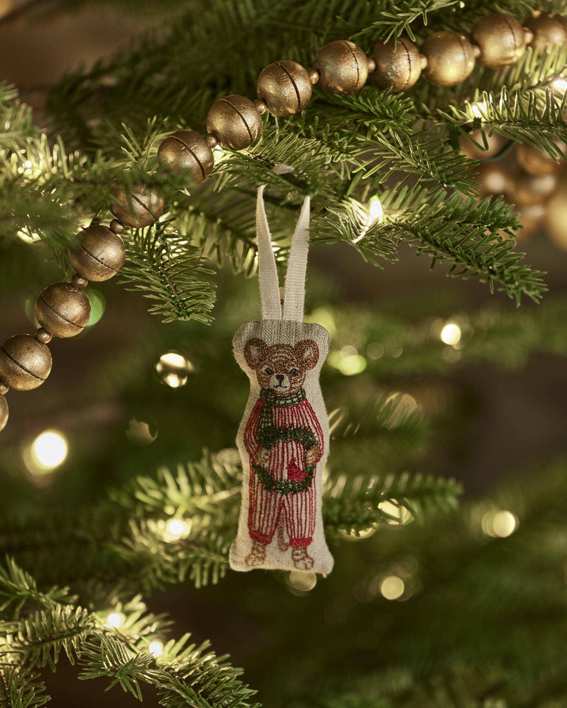 The Coral & Tusk Mouse with Wreath Ornament, dressed in red overalls and holding a wreath, hangs from a Christmas tree branch decorated with golden bead garland and glowing string lights.
