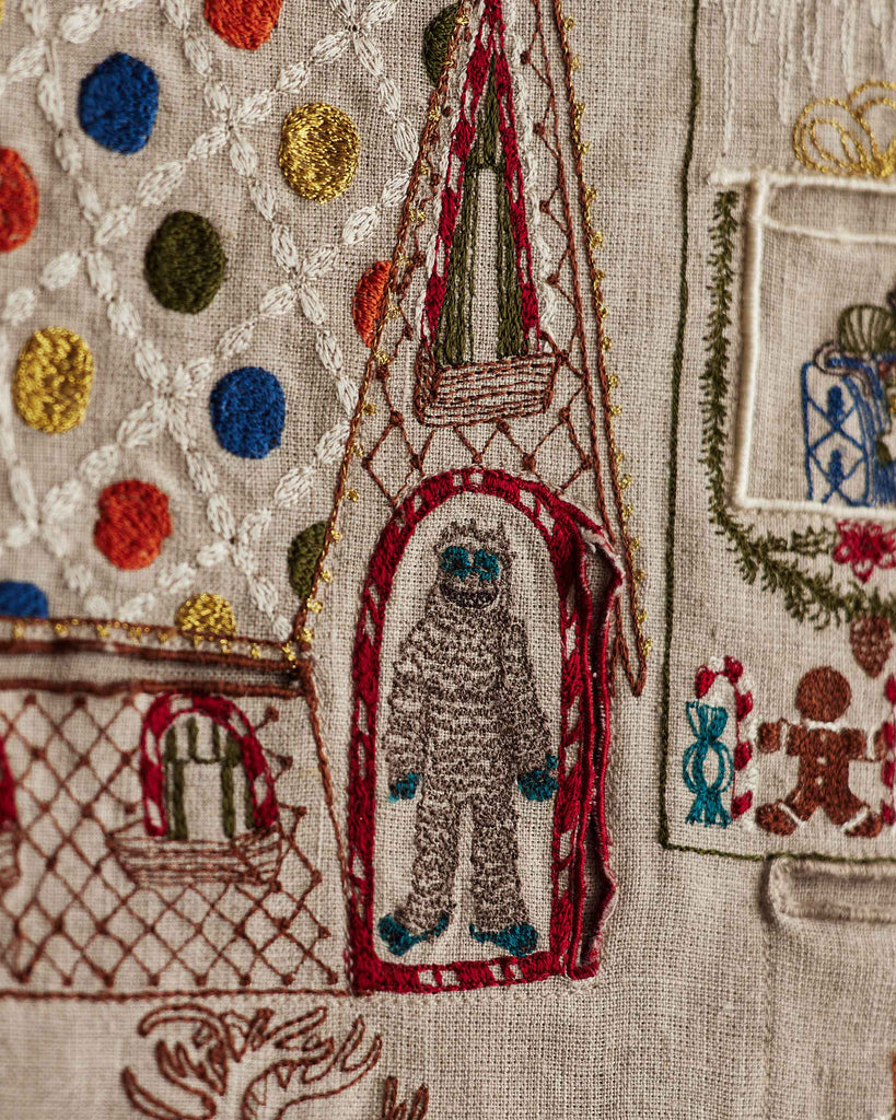 A close-up of the Coral & Tusk North Pole Advent Calendar shows colorful embroidery, including a small white figure with blue hands and feet in a pointed archway, alongside stitched polka dots, windows, gifts, gingerbread men, and other festive motifs.