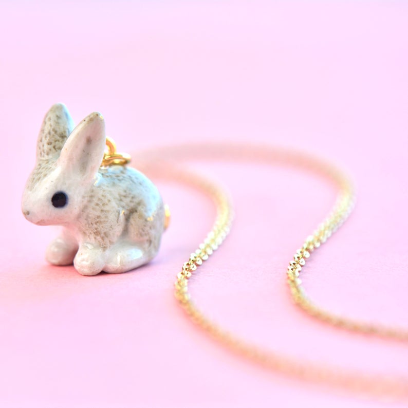 A delicate Goldentail Rabbit Necklace featuring a small, detailed hand-painted porcelain bunny pendant is laid out against a soft pink background. The chain is a fine 24k gold plated tone and loops gracefully around the bunny. The bunny pendant is white with gray speckled markings and a black eye.