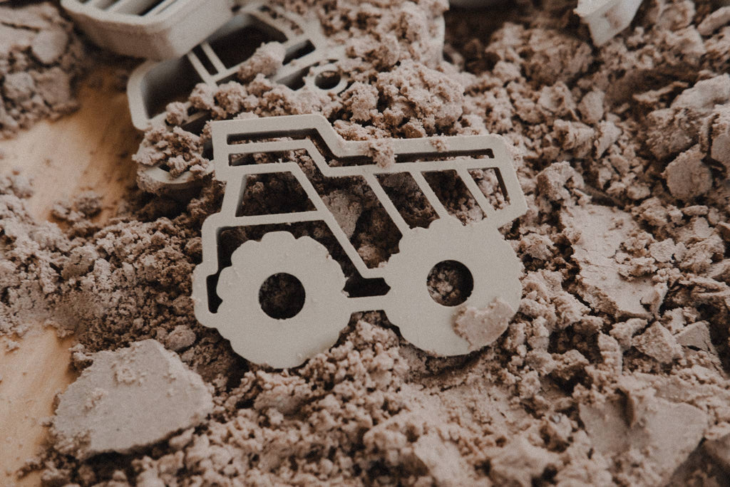 A Construction Cutter Set - Eco Playdough Tool shaped like a dump truck rests on a surface covered with brown kinetic sand, surrounded by scattered shapes and chunks—perfect for sensory play with your favorite playdough tools.