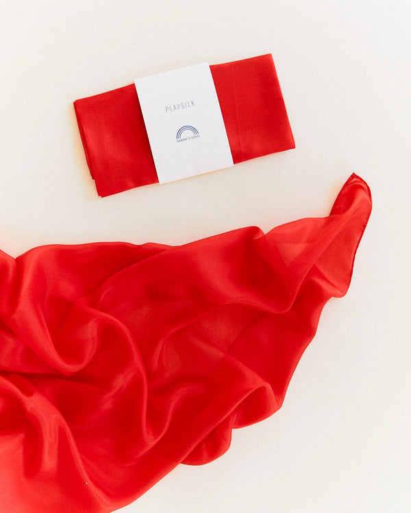 A folded Sarah's Silks Solid Color Playsilk - Red, made from pure silk and featuring a white label, rests above an unfurled red playsilk—ideal for open-ended play—against a light background.