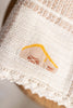 Close-up of the Organic Cotton Baby Blanket - Farm, featuring textured beige organic cotton with an embroidered house (yellow roof, brown windows), chickens, and a scalloped crochet trim at the bottom.