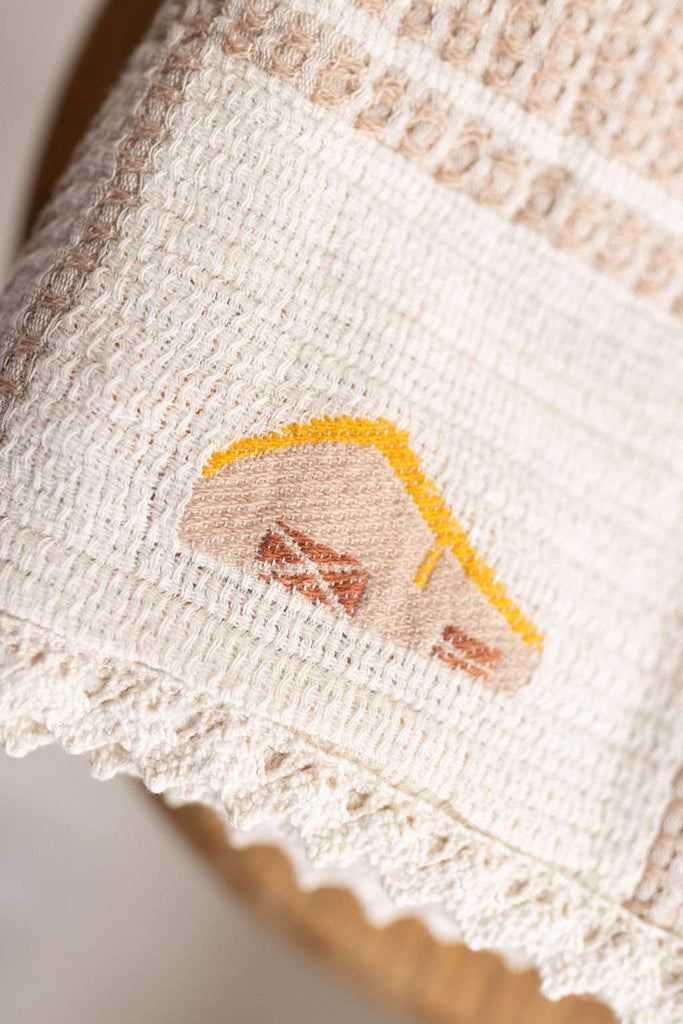 Close-up of the Organic Cotton Baby Blanket - Farm, featuring textured beige organic cotton with an embroidered house (yellow roof, brown windows), chickens, and a scalloped crochet trim at the bottom.
