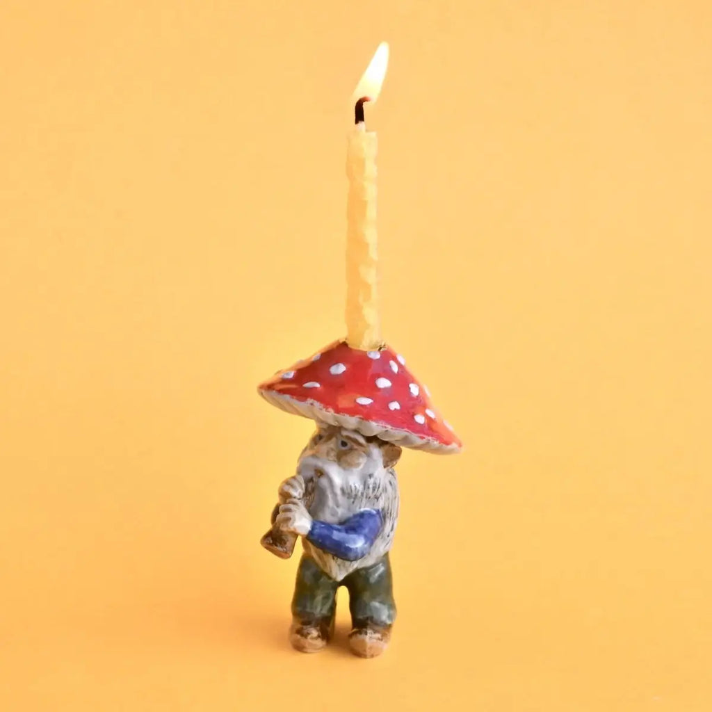 A whimsical Mushroom Gnome Cake Topper, hand painted, with a lit wick on top, set against a bright orange background.