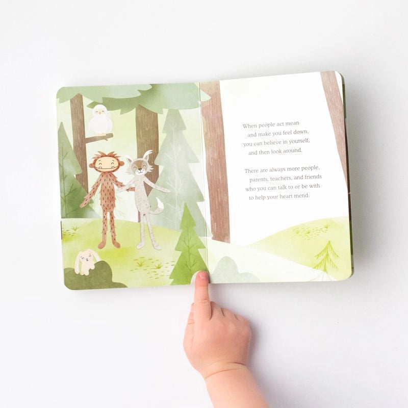 A child's hand pointing at a Slumberkins Bigfoot Snuggler + Intro Book - Self Esteem opened to a page showing a forest scene with a monkey, sheep, and a text about children's self-confidence and friendship.