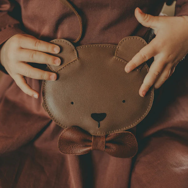 A child's hands holding a small, brown, Donsje Britta Exclusive Purse | Winter Bear-shaped purse with a cute bow tie, resting on their lap covered with a burgundy fabric.
