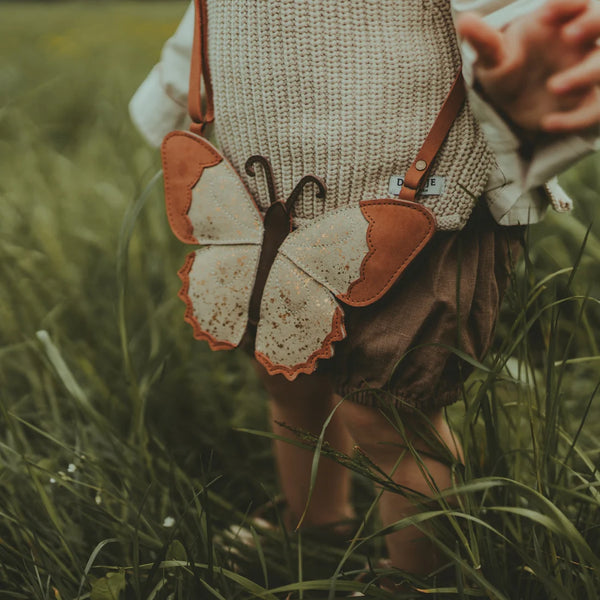 A toddler from behind holding a Donsje Toto Purse in a grassy field. The purse, featuring premium cream metallic suede with orange, black, and white colors resembling butterfly wings, includes an adjustable leather strap.