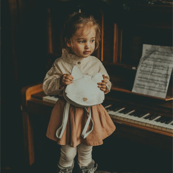 A young girl with a thoughtful expression holds a white plush heart and stands next to a piano with sheet music. She is dressed in a beige and pink dress, with matching socks and a Donsje Mini Leather Backpack - Cat.