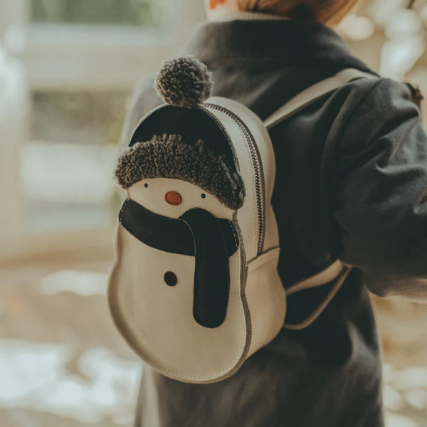 A child wearing a Donsje Kliff backpack with a snowman design, featuring a knit cap and cartoonish face, viewed from behind in a soft-focused, warm-lit setting.