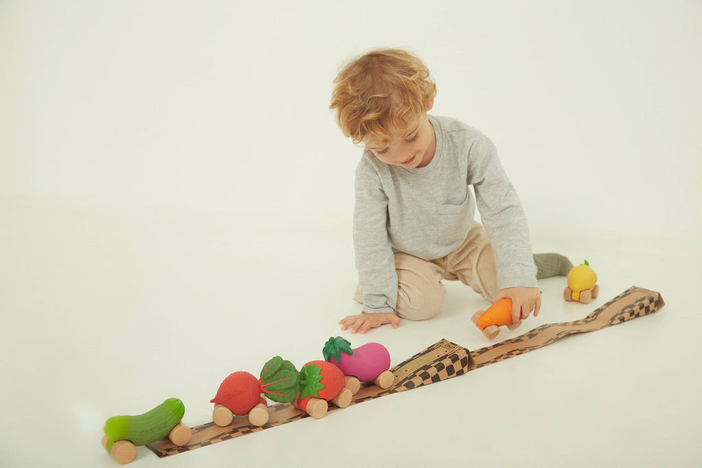 A young child sits on the floor, playing with Betty The Beetroot Baby Car, a colorful FSC-certified wood vegetable toy on wheels, lined up on a wooden track against a plain white background.