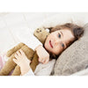 A young girl with blue eyes smiles on a bed, hugging the Senger Naturwelt Stuffed Animal - Beige Horse. She rests her head on a gray pillow and wears a white sweater.