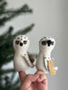 A hand holds two Snowy Owl Finger Puppets from the 2024 Finger Puppet Collection, featuring white bodies with black speckles. Each handmade puppet has a tag attached. A blurred evergreen tree is visible in the background.