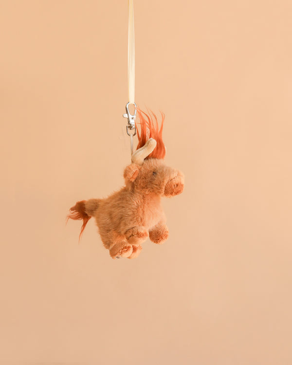 The Bukowski - Henderson Highland Cow Bag Charm, a small and light brown plush toy with an orange mane and tail, hangs from a white ribbon against a plain beige background, creating a whimsical accessory that brings charm and character to any bag.