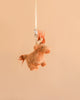 The Bukowski - Henderson Highland Cow Bag Charm, a small and light brown plush toy with an orange mane and tail, hangs from a white ribbon against a plain beige background, creating a whimsical accessory that brings charm and character to any bag.