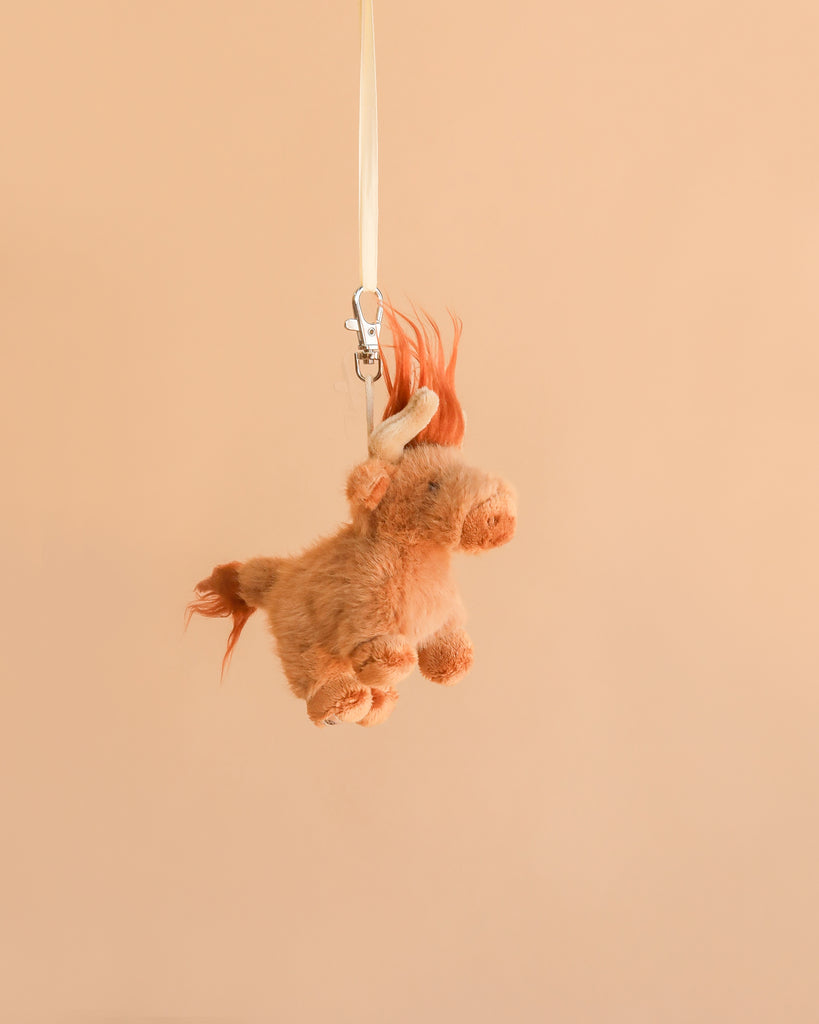 The Bukowski - Henderson Highland Cow Bag Charm, a small and light brown plush toy with an orange mane and tail, hangs from a white ribbon against a plain beige background, creating a whimsical accessory that brings charm and character to any bag.