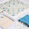 A Scrabble Bianco Edition board game featuring white and blue tiles on a wood-framed board, with a gold-topped hourglass, a blue tile bag, a score pad, and a pencil.