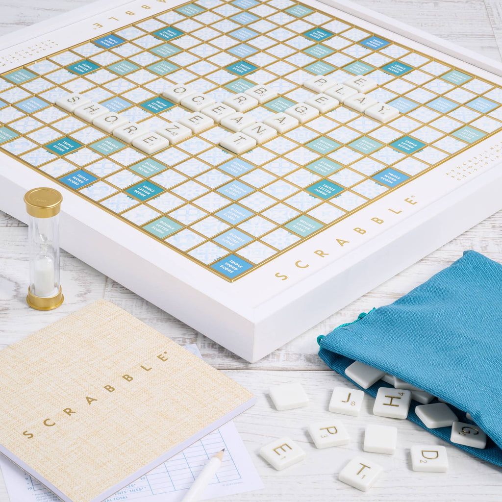 A Scrabble Bianco Edition board game featuring white and blue tiles on a wood-framed board, with a gold-topped hourglass, a blue tile bag, a score pad, and a pencil.