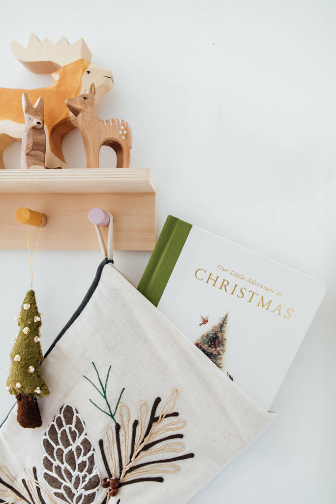 A Christmas stocking with pinecone embroidery hangs on a wooden hook. Nearby, “Our Little Adventure at Christmas (Baby’s First Christmas)” board book peeks out—perfect for toddler language skills and holiday gifting. Above, wooden animal figurines rest on a shelf.