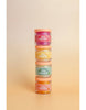 Four jars of "Tiny Tummies- Banana Jelly food - Tiny Harlow" are stacked vertically, each a different color: pink, orange, yellow, and green. The background is a soft beige gradient that subtly hints at the magic of feeding even the tiniest tummies with ease.