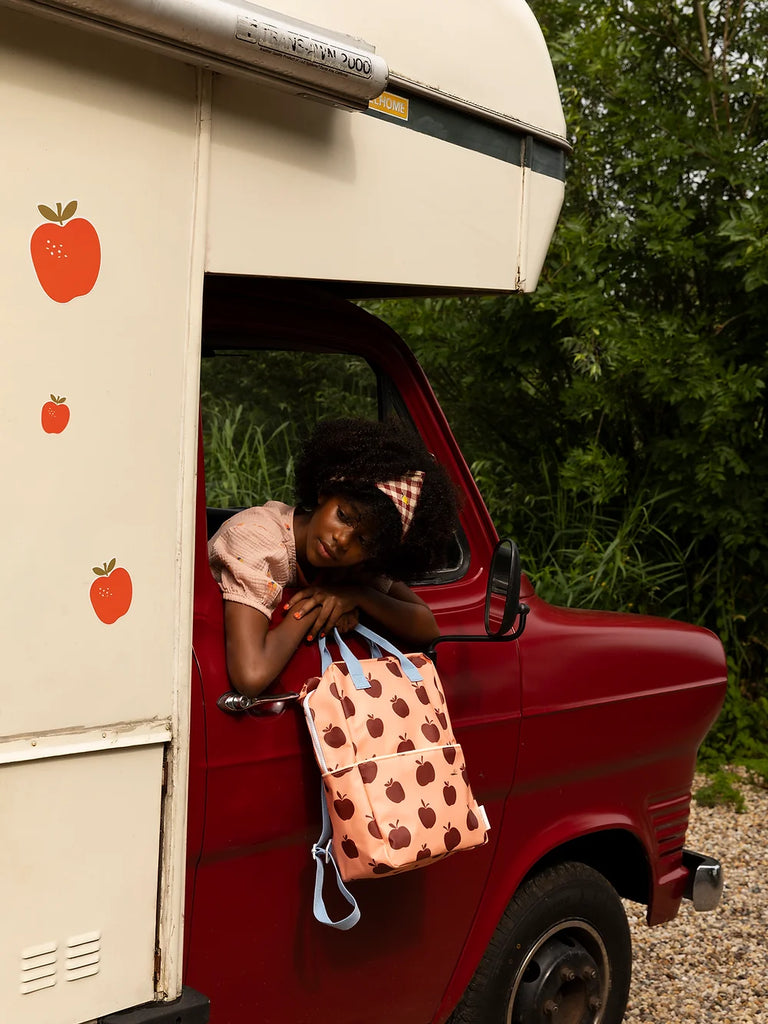 A young woman leans out of the window of a vintage red and white camper van, surrounded by lush greenery. She wears a pink blouse, a patterned headscarf, and holds a Sticky Lemon Backpack Large | Special Edition.