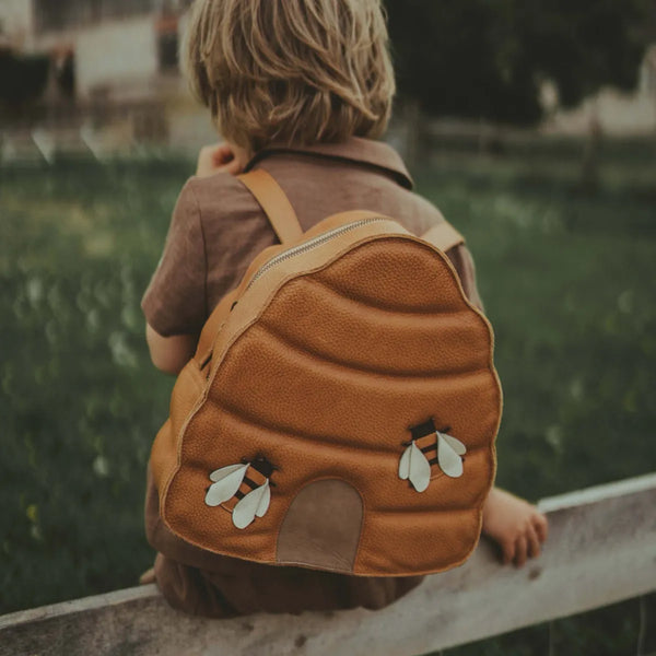 A young boy with a Donsje School Leather Backpack - Beehive shaped like a cute animal, sitting on a wooden fence in a lush green field. The focus is on his back, emphasizing a sense of