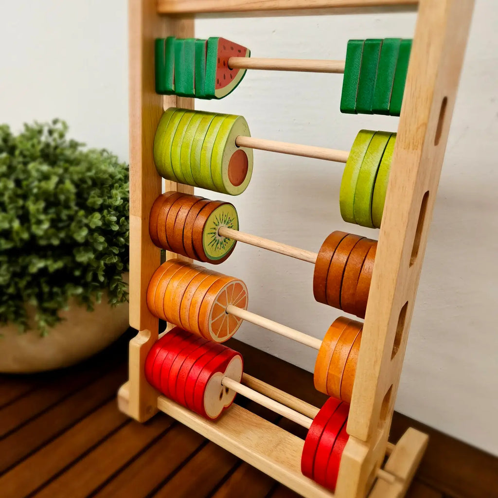 The Fruitful Abacus is a wooden toy featuring five rows of colorful, fruit-themed beads. These include a watermelon slice, two types of green rings, a kiwi slice, an orange slice, and an apple slice. This educational tool is perfect for enhancing counting skills. A potted green plant can be seen partially in the background.