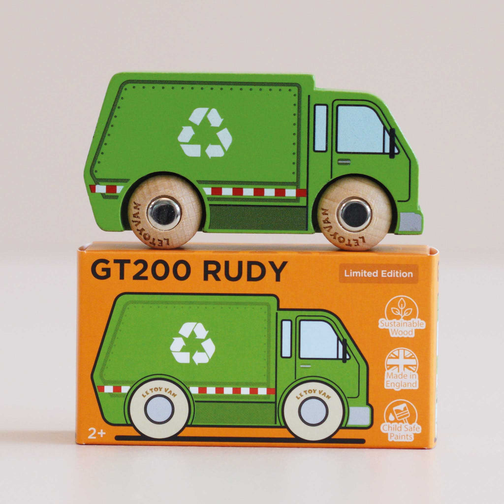 A green wooden recycling truck, labeled “No. 36 GT200 Rudy | Made in England,” sits atop an orange box adorned with white recycling symbols and red-striped accents. Both the truck and the box highlight eco-friendly toy features, inspiring sustainable play for young environmentalists.