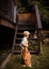 A young child in a beige cap and light outfit stands on grass, holding the Senger Naturwelt Cuddly Animal - Rust Goose, looking up at wooden stairs leading to a porch surrounded by greenery.